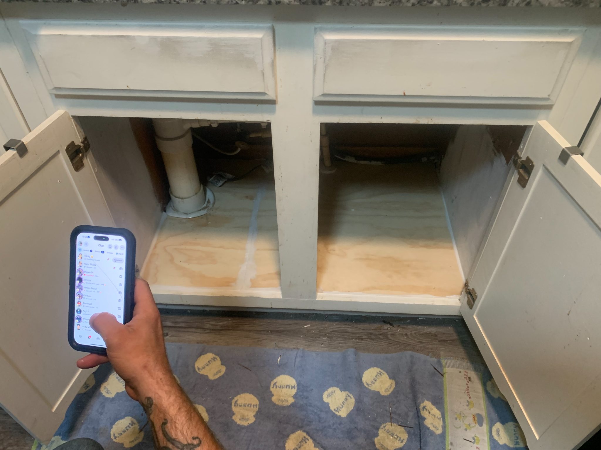 Person holding phone inside open kitchen cabinet with plumbing pipes visible