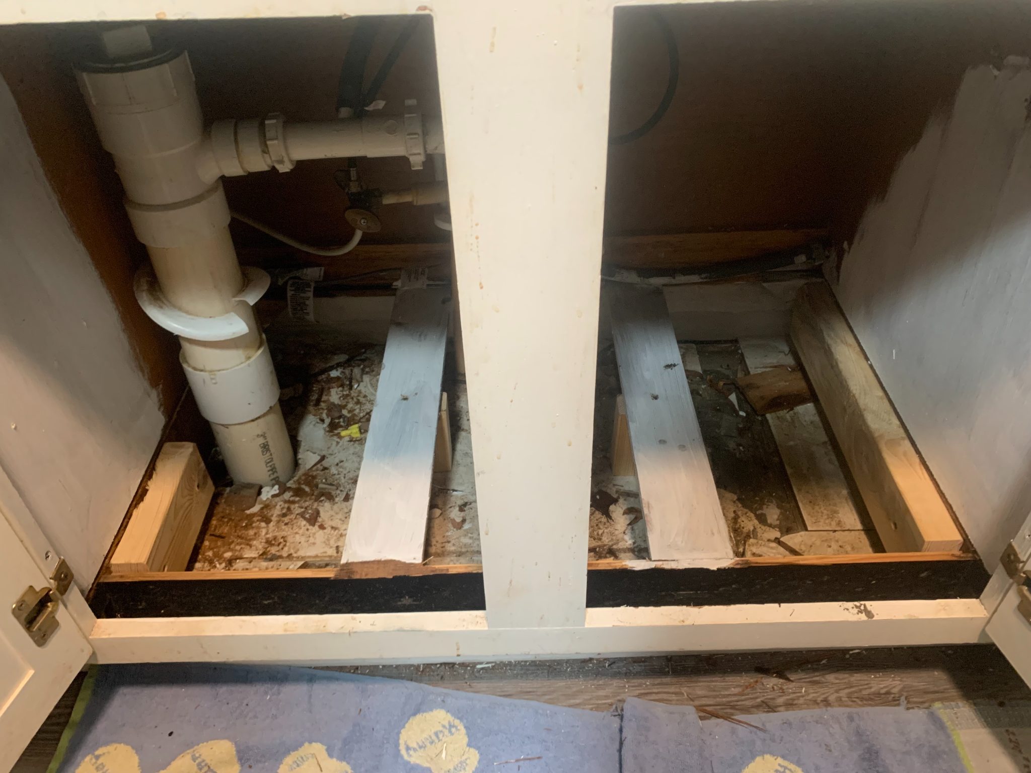 Interior view of bathroom vanity cabinet showing plumbing pipes and wooden framing with debris