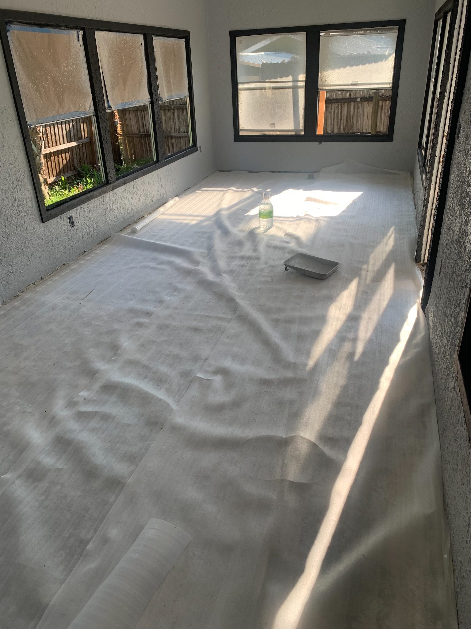 Empty room with concrete floor, white walls, black-framed windows, and protective plastic sheeting