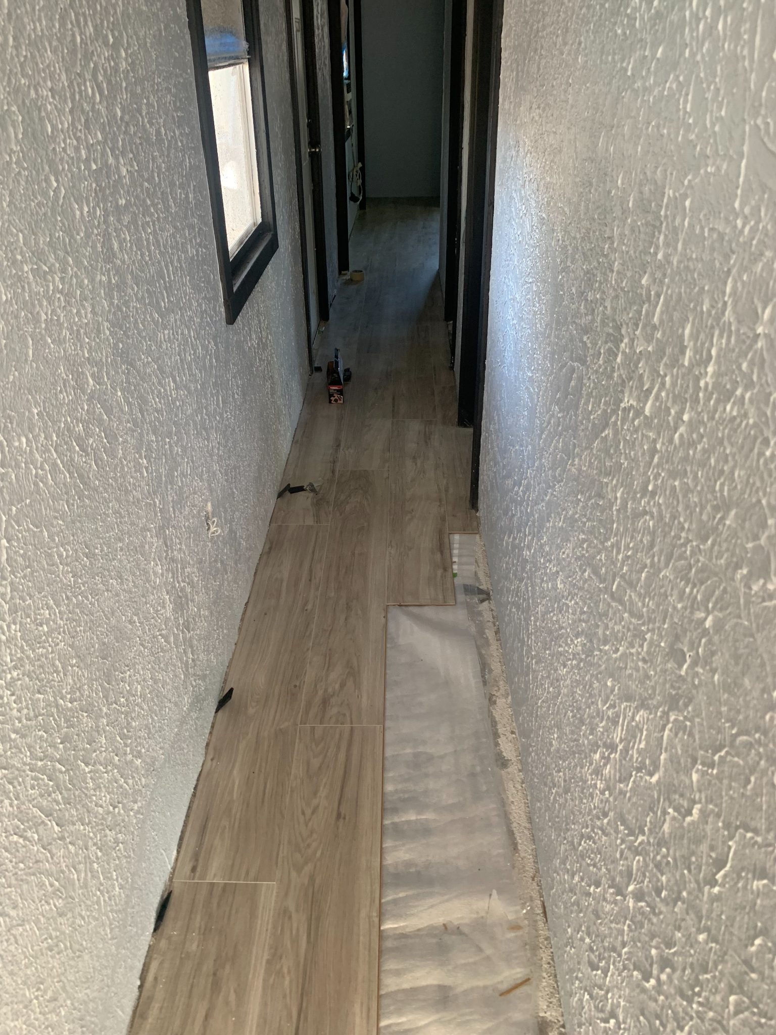 Narrow hallway with textured white walls, dark doors, wood-grain flooring, and natural light from a window